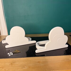 May include: Two white cloud-shaped shelves with a curved design, resting on a black surface. Several gold screws and small white plugs are next to the shelves. The background is a teal wall and a wooden table.