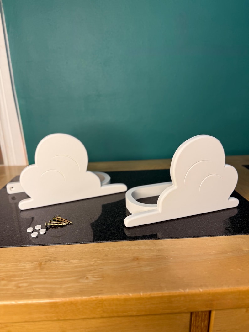 May include: Two white cloud-shaped shelves with a curved design, resting on a black surface. Several gold screws and small white plugs are next to the shelves. The background is a teal wall and a wooden table.