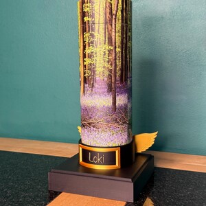 May include: A cylindrical container featuring a forest scene with purple bluebell flowers and tall trees. The base is black with a gold-colored plaque displaying the name Loki in black text. A small gold-colored wing extends from the side of the base.