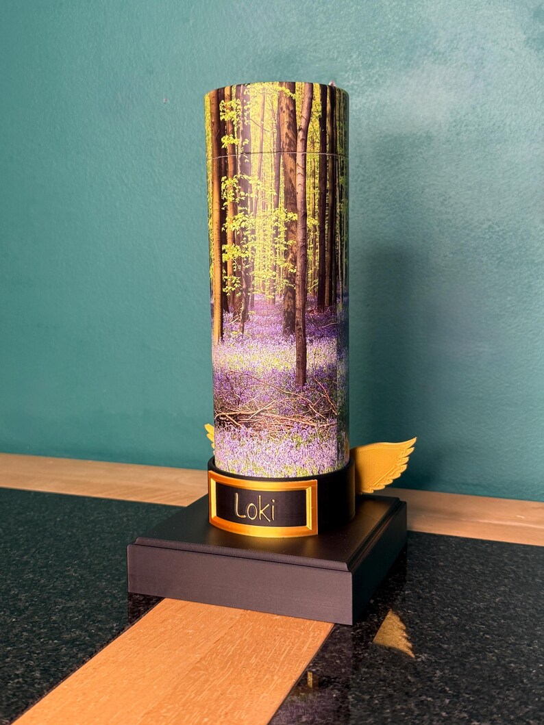 May include: A cylindrical container featuring a forest scene with purple bluebell flowers and tall trees. The base is black with a gold-colored plaque displaying the name Loki in black text. A small gold-colored wing extends from the side of the base.