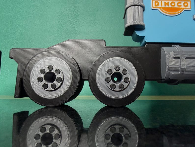 May include: A close-up of a toy truck's wheels and chassis. The wheels are black with gray centers and detailed bolt patterns. The truck's body is blue with the Dinoco logo visible. The image is well-lit, with a reflective surface below.