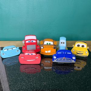 May include: A collection of colorful toy cars, including red, blue, yellow, and orange vehicles. The cars are arranged on a dark surface, with a teal background. The cars are cartoon-like and have distinct facial features.