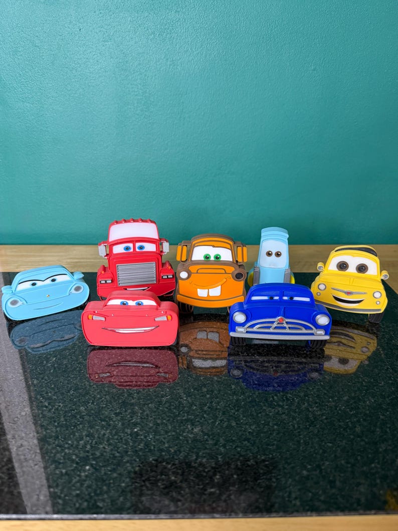May include: A collection of colorful toy cars, including red, blue, yellow, and orange vehicles. The cars are arranged on a dark surface, with a teal background. The cars are cartoon-like and have distinct facial features.