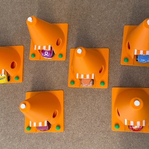 May include: An overhead shot of six orange 3D-printed traffic cones, each with a small toy car inside. The cones are on a square base with green accents. The toy cars are various colors and designs.