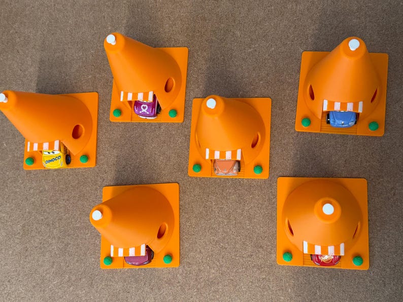 May include: An overhead shot of six orange 3D-printed traffic cones, each with a small toy car inside. The cones are on a square base with green accents. The toy cars are various colors and designs.