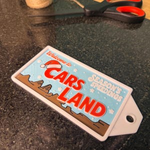 K&ouml;nnte beinhalten: Ein rechteckiges Schild mit dem Text "Welcome to Cars Land" in Rot und Wei&szlig;, mit einem Weihnachtsmannhut auf dem Text "Cars Land". Das Schild hat einen blauen Hintergrund mit braunen und wei&szlig;en Akzenten. Eine Schere und eine Schnurrolle befinden sich im Hintergrund.