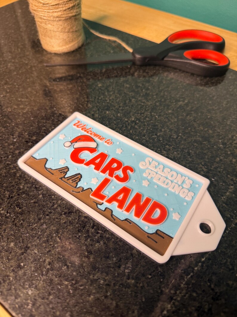 K&ouml;nnte beinhalten: Ein rechteckiges Schild mit dem Text "Welcome to Cars Land" in Rot und Wei&szlig;, mit einem Weihnachtsmannhut auf dem Text "Cars Land". Das Schild hat einen blauen Hintergrund mit braunen und wei&szlig;en Akzenten. Eine Schere und eine Schnurrolle befinden sich im Hintergrund.