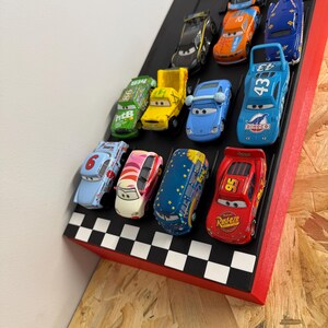 May include: A collection of toy race cars displayed on a black and red checkered platform. The cars are various colors, including blue, red, yellow, and green, and feature different racing numbers and designs. The platform is angled, suggesting a race track.
