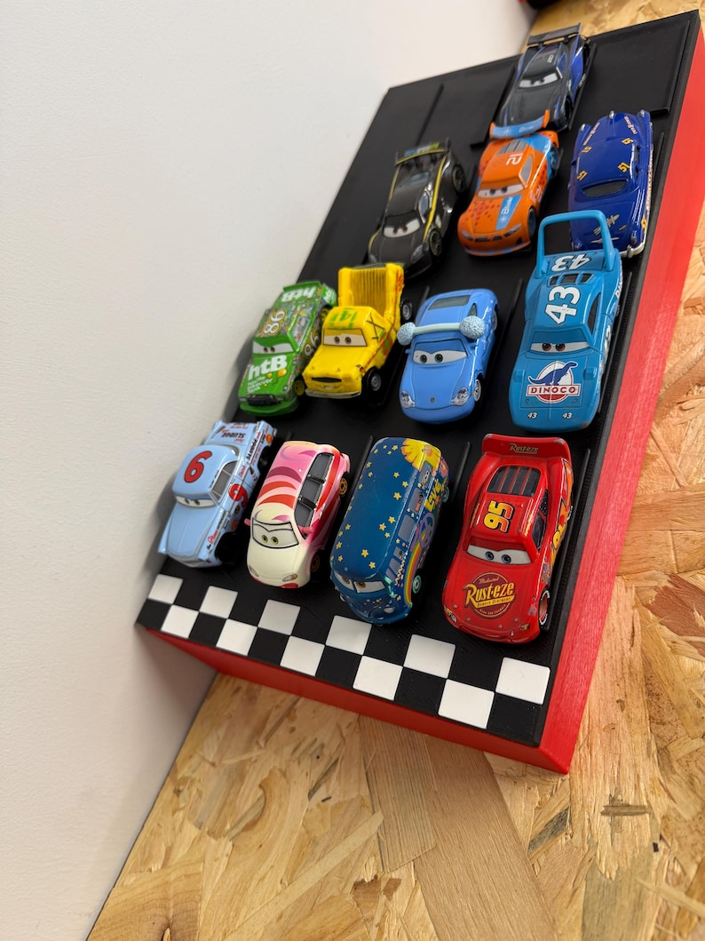 May include: A collection of toy race cars displayed on a black and red checkered platform. The cars are various colors, including blue, red, yellow, and green, and feature different racing numbers and designs. The platform is angled, suggesting a race track.