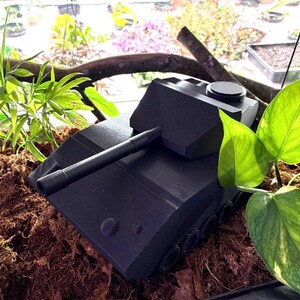 May include: A matte black toy tank model featuring a long barrel, a turret with a circular hatch, and visible side wheels. The tank is positioned among brown soil and green foliage, showcasing a geometric design with sharp angles and a solid structure.