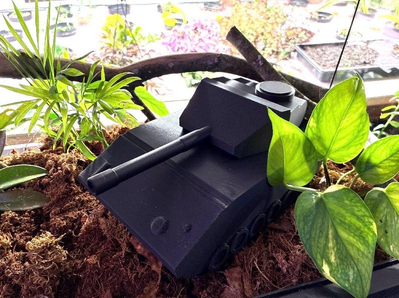 May include: A matte black toy tank model featuring a long barrel, a turret with a circular hatch, and visible side wheels. The tank is positioned among brown soil and green foliage, showcasing a geometric design with sharp angles and a solid structure.