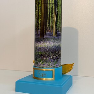 May include: A cylindrical container featuring a forest scene with purple flowers, mounted on a bright blue square base. A gold-colored plaque on the front displays the name Cain in script, and small gold-colored wings are attached to the sides.