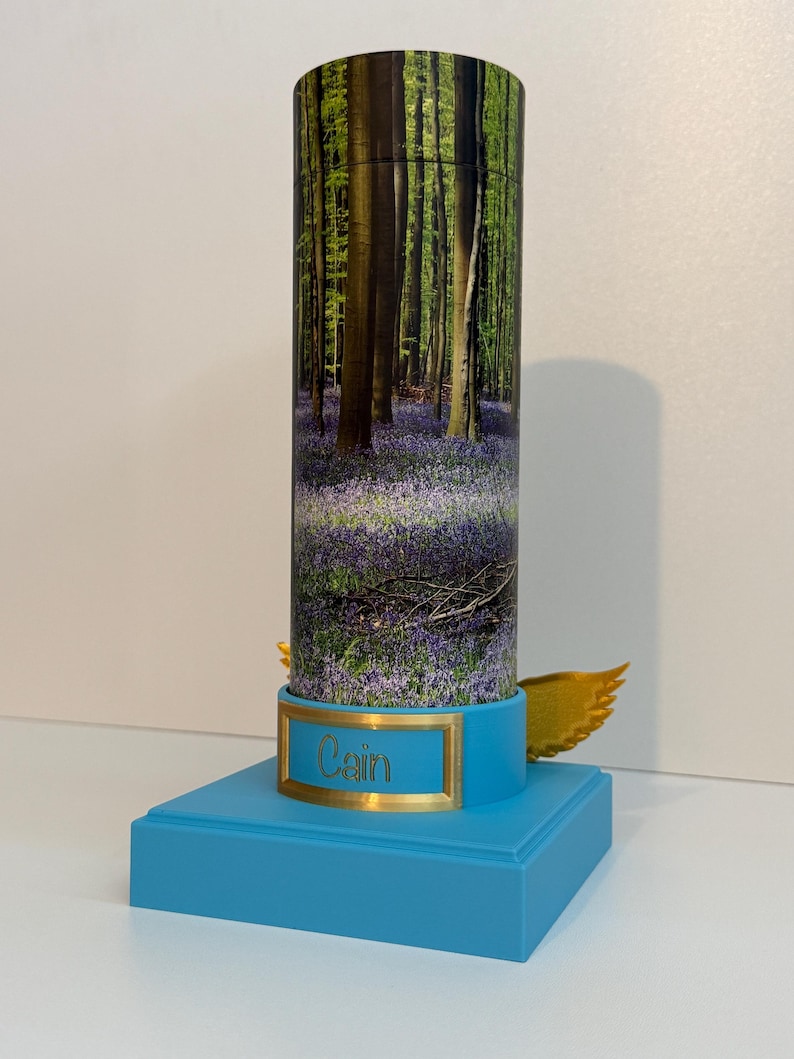 May include: A cylindrical container featuring a forest scene with purple flowers, mounted on a bright blue square base. A gold-colored plaque on the front displays the name Cain in script, and small gold-colored wings are attached to the sides.