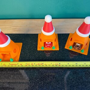 May include: Three orange traffic cones with red and white Santa hats, each housing a toy car. A yellow tape measure shows the length of the objects. The cones are on a black surface.