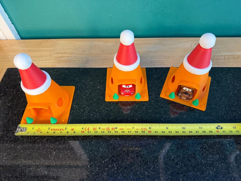 May include: Three orange traffic cones with red and white Santa hats, each housing a toy car. A yellow tape measure shows the length of the objects. The cones are on a black surface.