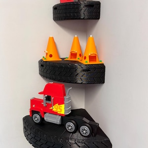 May include: Three-tiered corner shelf unit made from black tires. The top shelf holds toy cars, the middle shelf has orange traffic cones, and the bottom shelf displays a red toy truck. The shelves are mounted on a white wall.