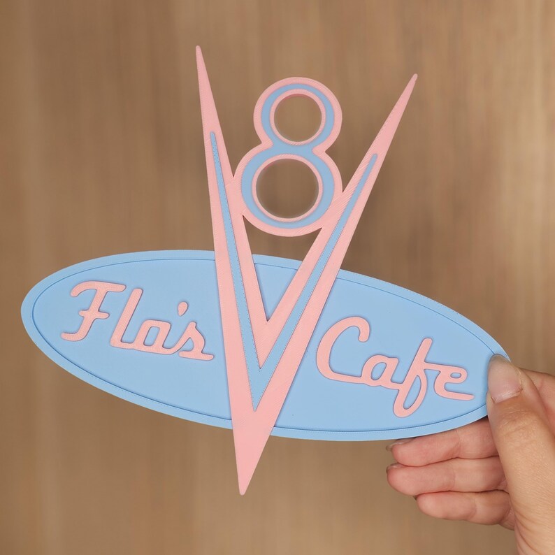 May include: A retro-style sign with a light blue oval base and pink lettering that reads "Flo's Cafe." A pink and light blue V-shaped design with an "8" in the center is above the oval. The sign is held by a hand.
