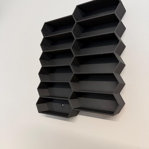 May include: A matte black wall-mounted display shelf featuring a modular honeycomb-inspired design with twelve individual rectangular compartments arranged in two vertical columns, creating a geometric storage structure for small items.