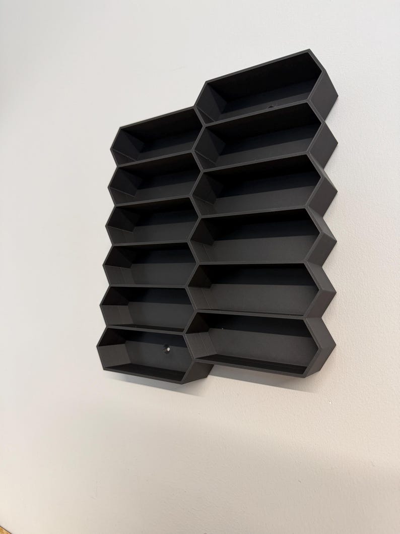 May include: A matte black wall-mounted display shelf featuring a modular honeycomb-inspired design with twelve individual rectangular compartments arranged in two vertical columns, creating a geometric storage structure for small items.