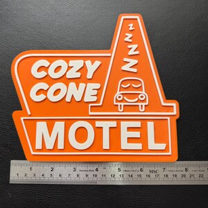 以下が含まれることがあります： オレンジと白の看板に「Cozy Cone Motel」の文字と、ZZZが書かれた漫画の車が描かれています。