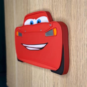 K&ouml;nnte beinhalten: Rote Lightning McQueen-Autodekoration mit einem l&auml;chelnden Gesicht, blauen Augen und wei&szlig;en Z&auml;hnen. Das Auto hat schwarze Reifen und orangefarbene und graue Details. Die Dekoration ist auf einer Holzoberfl&auml;che befestigt.