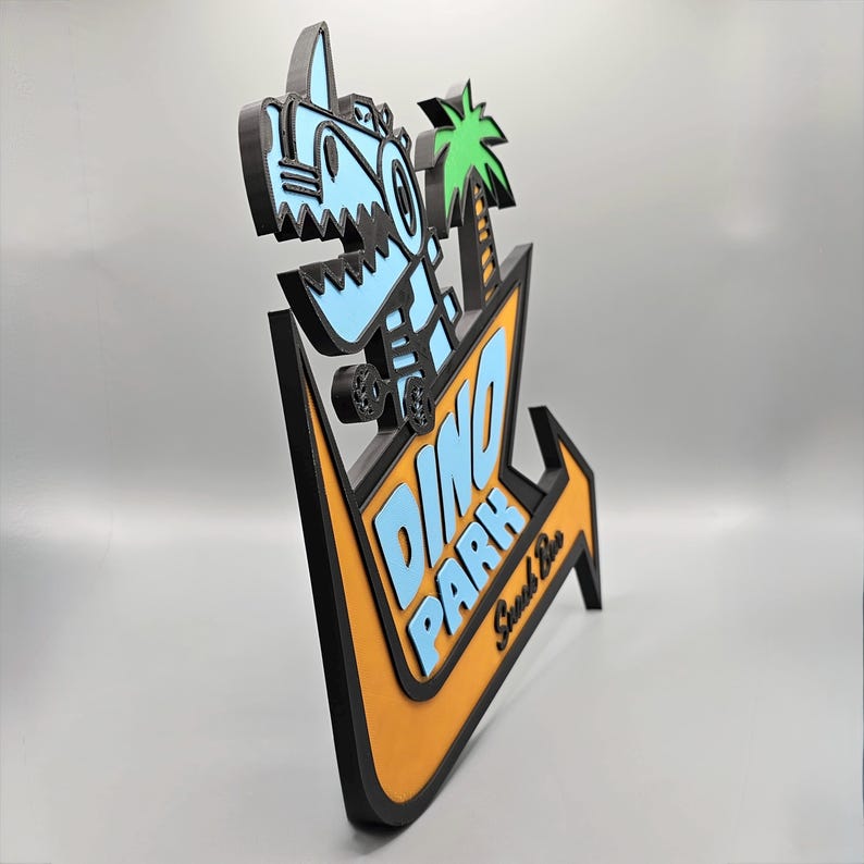 May include: A 3D-printed sign featuring a cartoon dinosaur and palm tree. The sign is in the shape of an arrow, with the words "DINO PARK" in blue and orange. The sign is black, blue, orange, and green.
