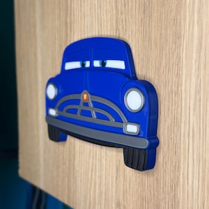 Pode incluir: Decora&ccedil;&atilde;o de parede em forma de carro azul com far&oacute;is e olhos brancos. O carro tem uma grelha e p&aacute;ra-choques cinzentos e pneus pretos. A decora&ccedil;&atilde;o &eacute; montada numa superf&iacute;cie de madeira clara.