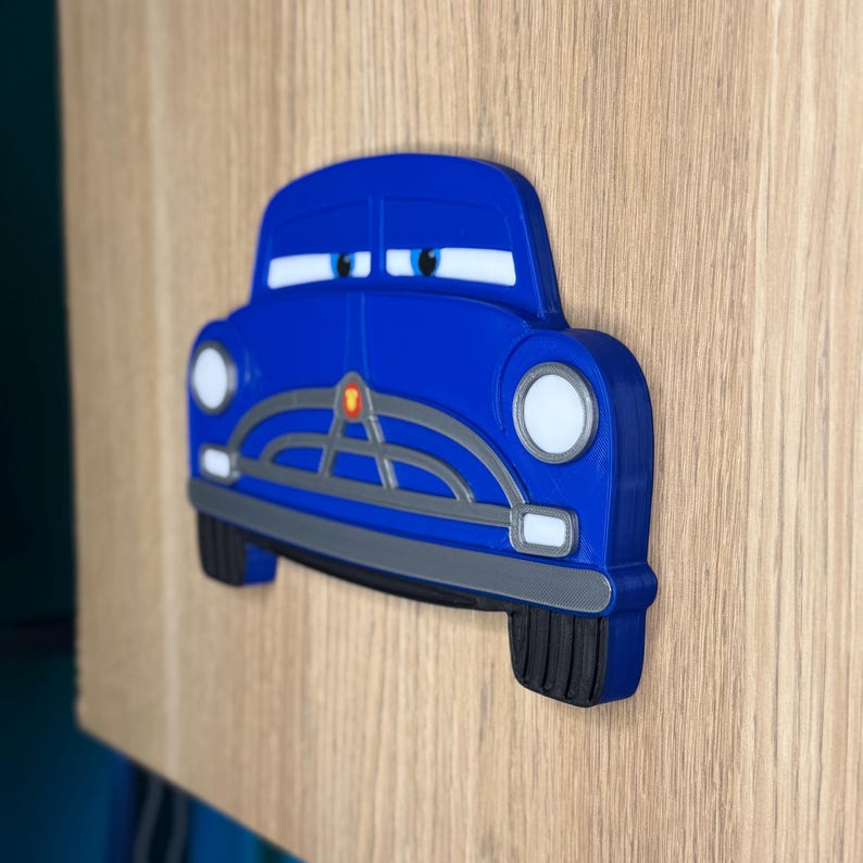 Pode incluir: Decora&ccedil;&atilde;o de parede em forma de carro azul com far&oacute;is e olhos brancos. O carro tem uma grelha e p&aacute;ra-choques cinzentos e pneus pretos. A decora&ccedil;&atilde;o &eacute; montada numa superf&iacute;cie de madeira clara.