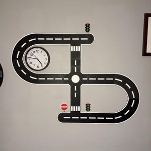 May include: A wall-mounted road play set with a clock, stop sign, and traffic lights. The black road pieces have white dashed lines and are arranged in a unique pattern. The clock has black hands and white numbers. The stop sign is red and white.
