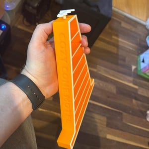 Op de afbeelding: Een oranje en wit Cozy Cone Motel model. Het model wordt in de hand van een persoon gehouden. Het model heeft de woorden "COZY CONE MOTEL" op de zijkant gedrukt. Het model heeft een witte structuur bovenop.