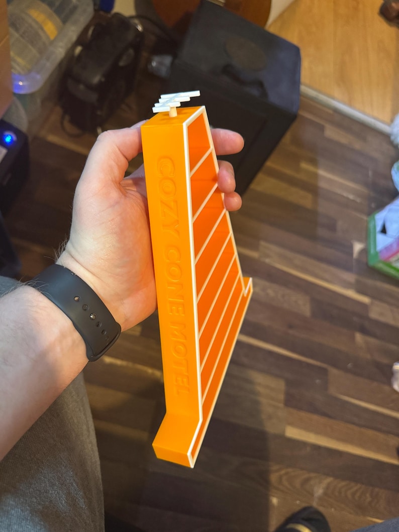 Op de afbeelding: Een oranje en wit Cozy Cone Motel model. Het model wordt in de hand van een persoon gehouden. Het model heeft de woorden "COZY CONE MOTEL" op de zijkant gedrukt. Het model heeft een witte structuur bovenop.