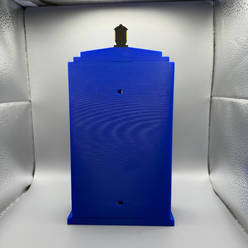 May include: A blue rectangular box with a black top, featuring two circular cutouts. The box has a textured surface and a stepped design. The object is a replica of a police call box.