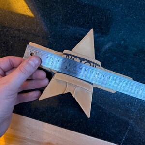 Puede incluir: Un objeto en forma de estrella de color beige con la palabra "VICTORY" impresa, medido con una regla. La estrella mide aproximadamente 20 cm de di&aacute;metro. El objeto est&aacute; sobre una superficie oscura.