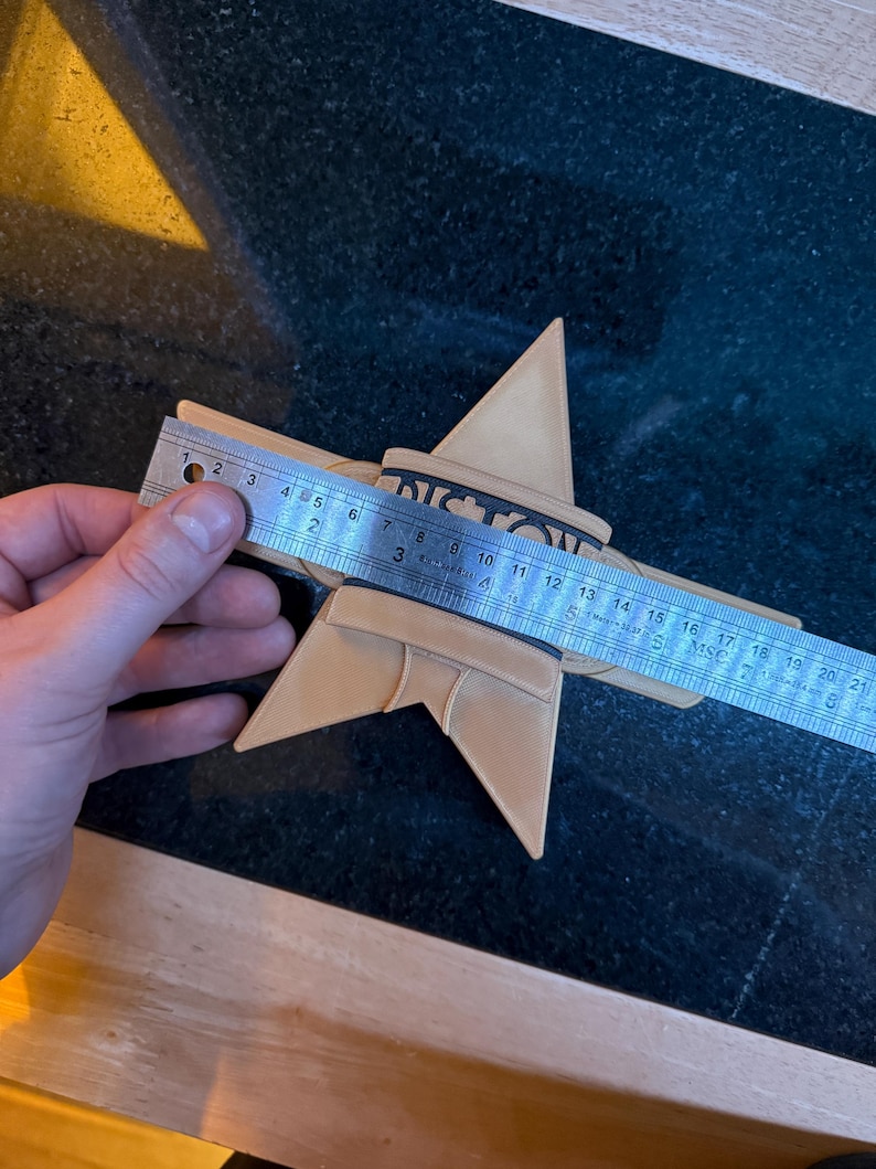 Puede incluir: Un objeto en forma de estrella de color beige con la palabra "VICTORY" impresa, medido con una regla. La estrella mide aproximadamente 20 cm de di&aacute;metro. El objeto est&aacute; sobre una superficie oscura.