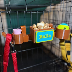 3D-printed PETG dog crate organiser in brown with a personalised nameplate. Holds a tennis ball, treats, poo bag roll, and two leashes. Heat-resistant for cars and vans, with adjustable hooks for easy attachment. Ideal for dog walkers and pet owners.