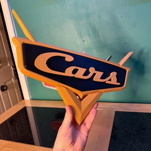 May include: A 3D-printed sign featuring the word "Cars" in orange lettering on a black background. The sign has a stylized design with orange accents and a triangular base. The sign is held up against a teal wall.