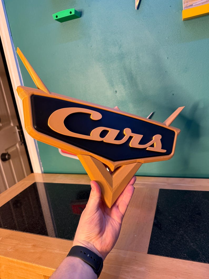 May include: A 3D-printed sign featuring the word "Cars" in orange lettering on a black background. The sign has a stylized design with orange accents and a triangular base. The sign is held up against a teal wall.
