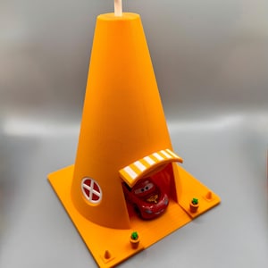 Op de afbeelding: Een feloranje speelhuis in de vorm van een verkeerskegel met een rode speelgoedauto erin. De kegel heeft een witte antenne bovenop en een wit afdak boven de ingang. Aan weerszijden van de ingang staan kleine groene plantjes.