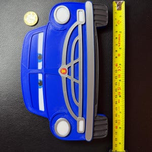 Pode incluir: Um carro de brinquedo azul com detalhes brancos e prateados, com olhos de desenho animado e uma grelha dianteira detalhada. O carro tem cerca de 23 cm de comprimento, com uma fita m&eacute;trica amarela ao lado. H&aacute; tamb&eacute;m uma moeda de ouro.