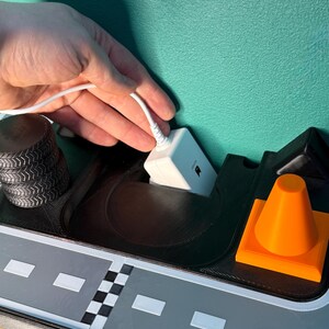 May include: A 3D-printed toy road scene featuring a grey road, black tyre stacks, an orange traffic cone, and a white electronic device with a cable. The scene is set against a teal background.