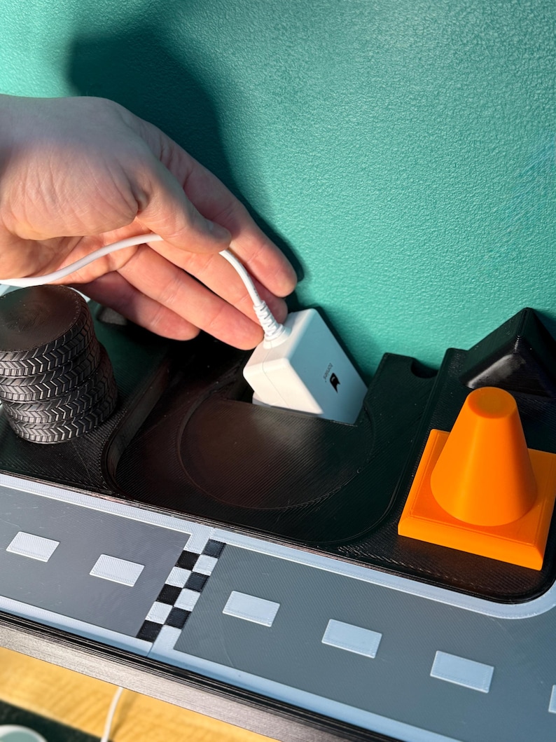 May include: A 3D-printed toy road scene featuring a grey road, black tyre stacks, an orange traffic cone, and a white electronic device with a cable. The scene is set against a teal background.