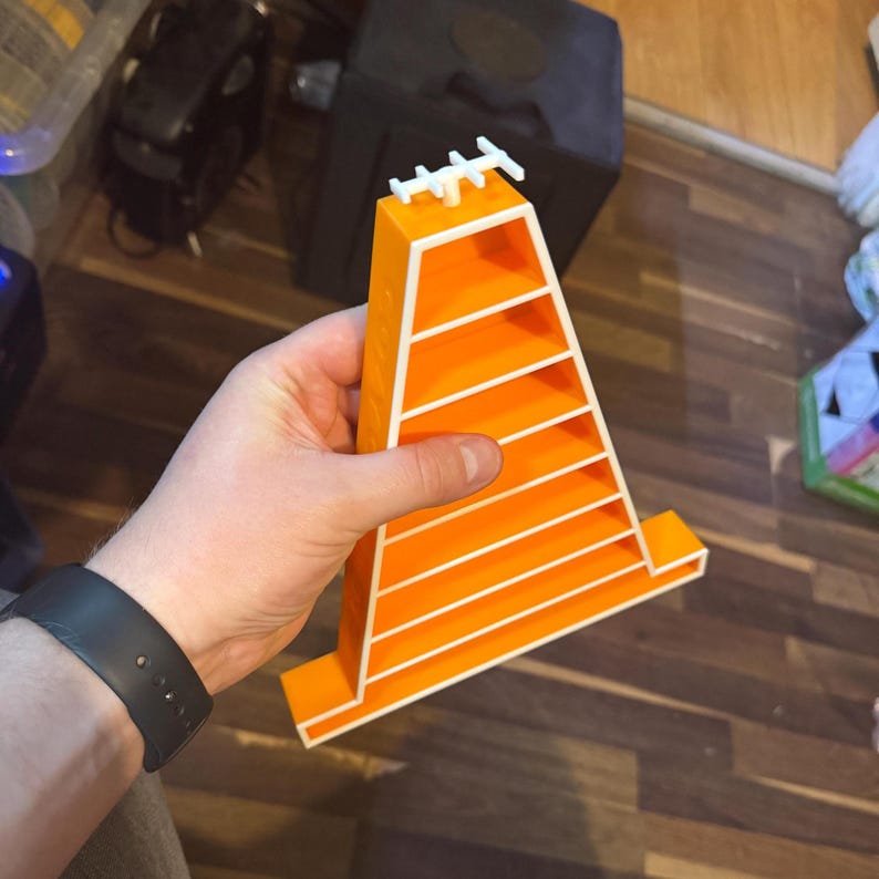 Op de afbeelding: Een oranje en witte gelaagde displaystandaard, vastgehouden in een hand. De standaard heeft een driehoekige vorm met horizontale planken en een decoratieve bovenkant. De standaard is gemaakt van plastic.
