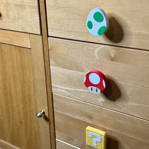 May include: Wooden cabinet with three decorative knobs featuring iconic designs: a white egg with green spots, a red mushroom with a white face, and a yellow question mark block. The knobs are attached to the cabinet drawers.