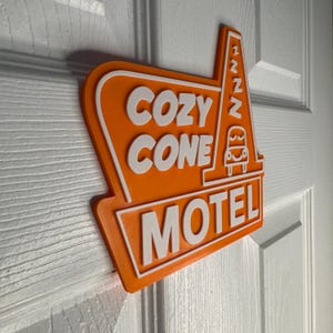 以下が含まれることがあります： オレンジと白の看板に、白い文字で「Cozy Cone Motel」と書かれています。看板には、車のスタイリッシュな画像と、側面に「zzz」という文字があります。