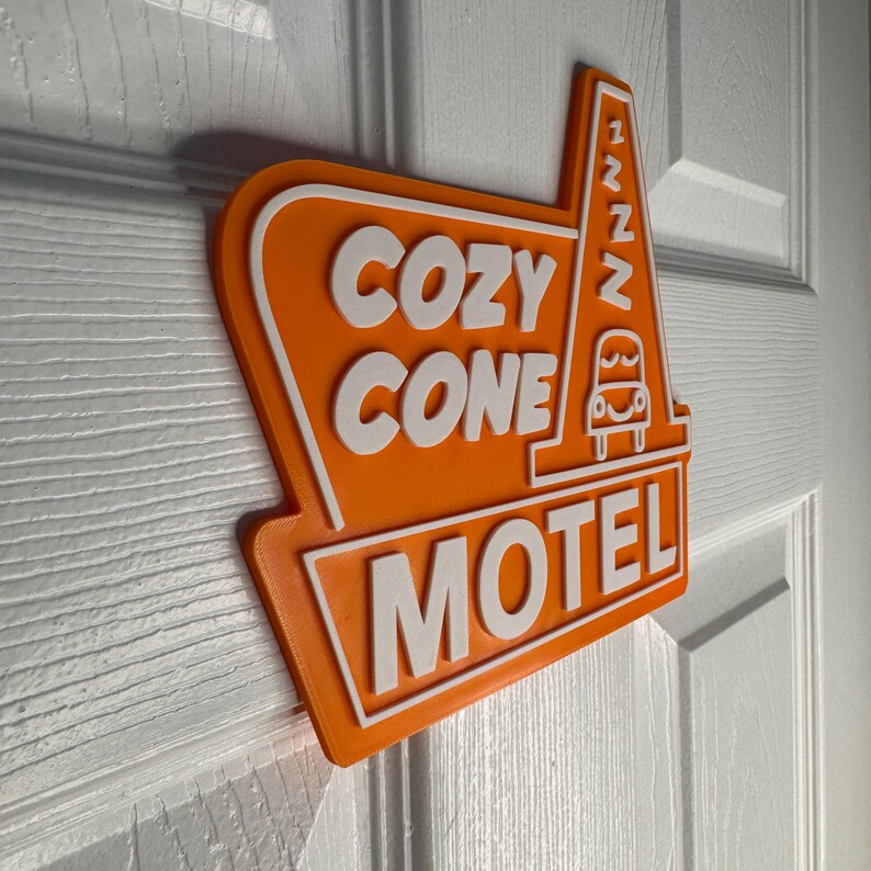 以下が含まれることがあります： オレンジと白の看板に、白い文字で「Cozy Cone Motel」と書かれています。看板には、車のスタイリッシュな画像と、側面に「zzz」という文字があります。
