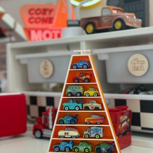 Op de afbeelding: Een oranje en witte displayplank in de vorm van een driehoek, gevuld met miniatuur speelgoedauto's in verschillende kleuren. De plank staat op een witte plank met andere speelgoedauto's en een bord met "Cozy Cone Motel".
