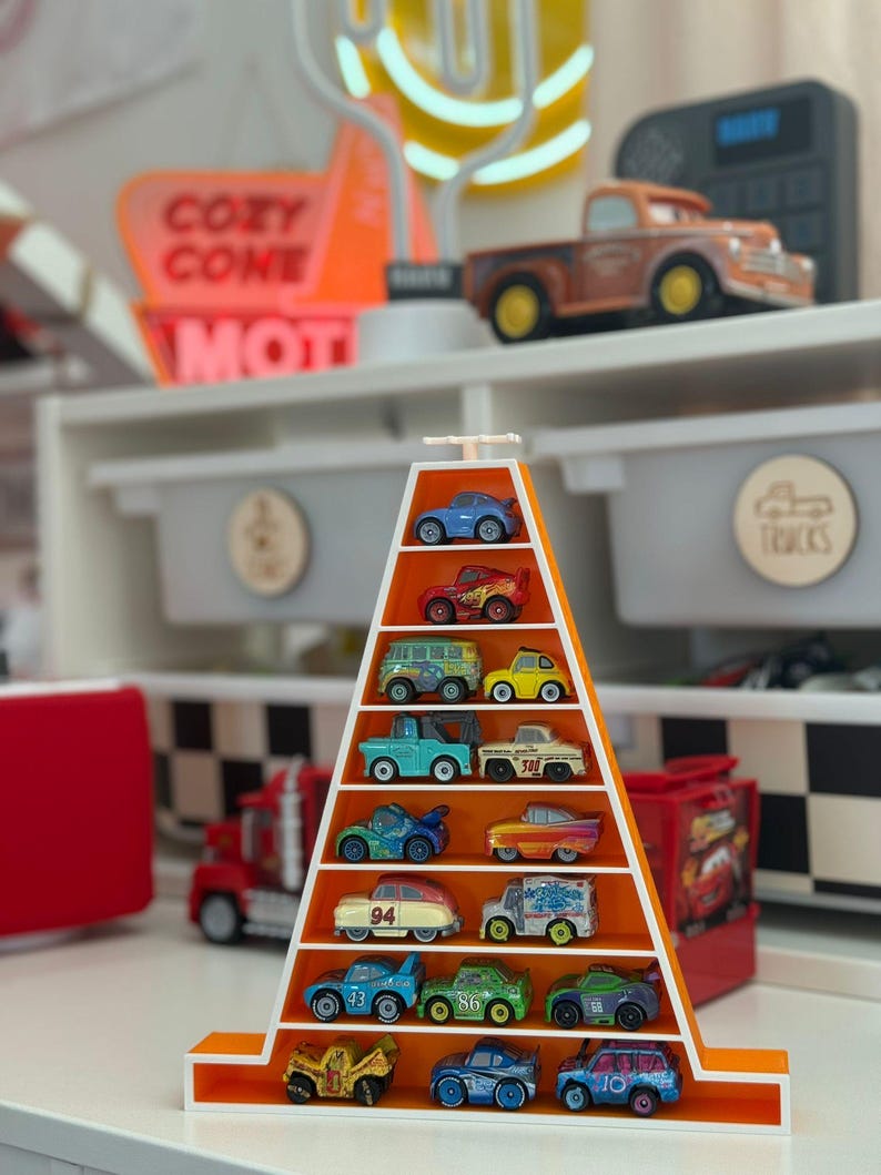 Op de afbeelding: Een oranje en witte displayplank in de vorm van een driehoek, gevuld met miniatuur speelgoedauto's in verschillende kleuren. De plank staat op een witte plank met andere speelgoedauto's en een bord met "Cozy Cone Motel".