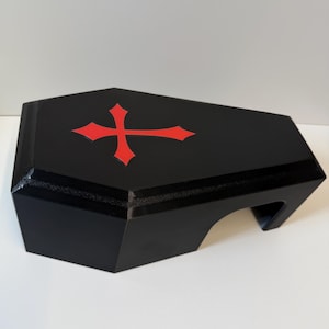 Könnte beinhalten: Eine schwarze, sargförmige Box mit glatter Oberfläche und einem roten Kreuzemblem in der Mitte des Deckels. Das Objekt hat eine hexagonale Silhouette mit einer flachen Oberseite und einem ausgeschnittenen Bereich an der Seite.