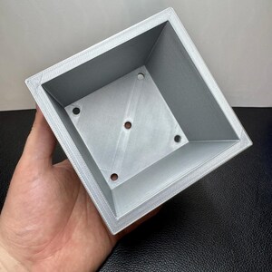 May include: A square, light gray planter with a textured surface and four drainage holes. The planter has a beveled edge and is held in a hand against a neutral background. The planter is designed for small plants.