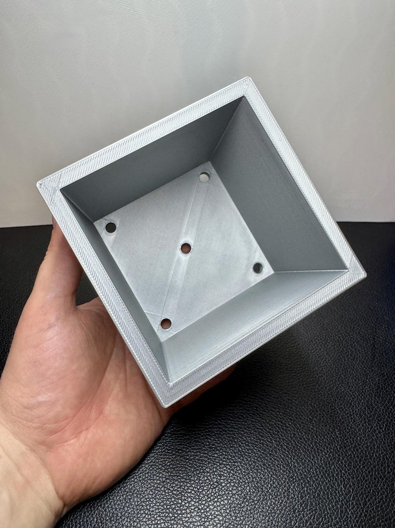 May include: A square, light gray planter with a textured surface and four drainage holes. The planter has a beveled edge and is held in a hand against a neutral background. The planter is designed for small plants.